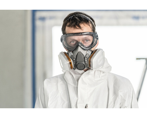 Een persoon draagt een veiligheidsbril, een ademhalingsmasker en beschermende kleding.