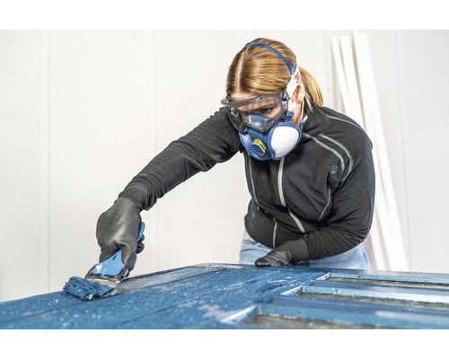 Vrouw met veiligheidsbril en ademhalingsmasker schildert deur met penseel