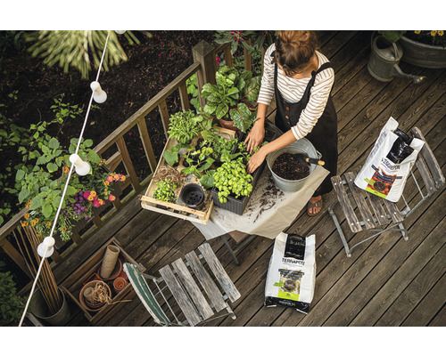 Vrouw verpot planten op een houten terras met potgrond, omringd door tuingereedschap en decoratie