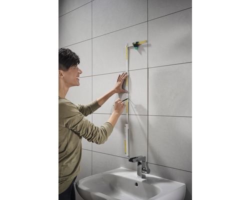 Vrouw meet een betegelde muur met een duimstok en een potlood in de badkamer boven een wastafel.