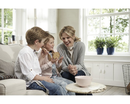 Een moeder en haar kinderen zitten op een bank en eten koekjes in een lichte woonkamer.
