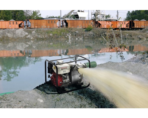 Waterpomp in werking naast een waterpartij met treinwagons op de achtergrond.