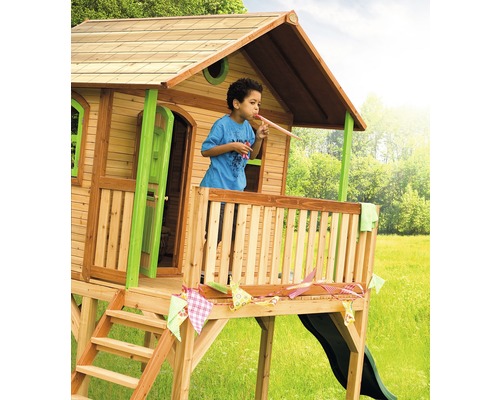 Houten speelhuis met glijbaan en een kind op de veranda dat in een feesttoeter blaast