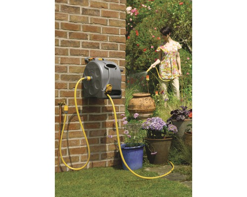 Slanghaspel met gele tuinslang gemonteerd op een bakstenen muur. Een vrouw geeft planten water met de slang.