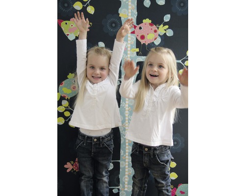 Twee meisjes staan voor een kindermeetlat met vogelmotieven op een donkere muur met bloemmotieven.