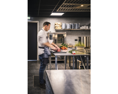 Een kok bereidt groenten en vlees op een roestvrijstalen tafel in een professionele keuken.