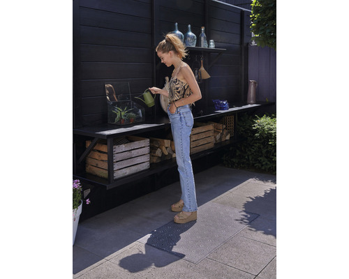 Een vrouw geeft planten water met een gieter op een terras met planken, houten kisten en een vloermat.