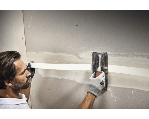 Een man bewerkt een gipsplaatwand met een plamuurmes en een wapeningsband.