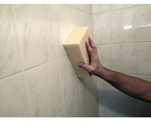 Een hand houdt een spons tegen een betegelde muur.