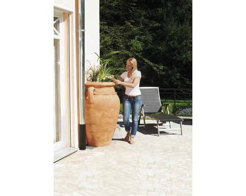 Vrouw geeft planten water in een terracotta plantenbak op een terras.