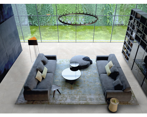 Lichte woonkamer met tegelvloer, banken, tafel en boekenkast voor een groot raam.