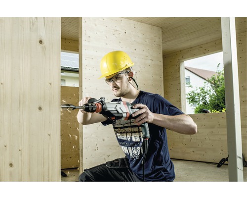 Een man boort met een boormachine een gat in een houten plank. Hij draagt een veiligheidshelm en een veiligheidsbril.