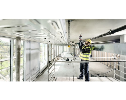 Een bouwvakker op een steiger werkt met een boorhamer aan een plafondinstallatie.