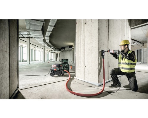 Een bouwvakker boort met een boorhamer een gat in een betonnen muur, stofafzuiging is aangesloten.