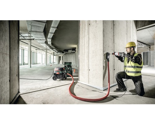 Een bouwvakker boort met een boormachine een gat in een betonnen muur. Een industriële stofzuiger en een afzuigadapter zijn ook aanwezig.