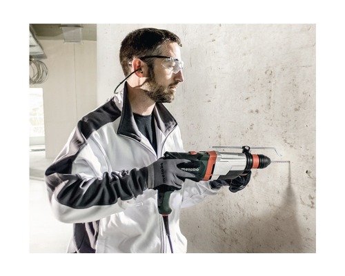 Een man boort met een Metabo boorhamer een gat in een betonnen muur. Hij draagt een veiligheidsbril, oorbeschermers en handschoenen.