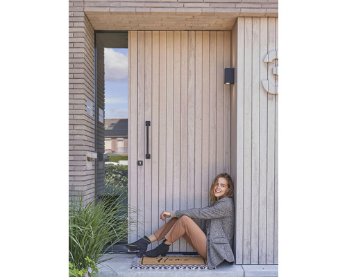 Vrouw zit voor een voordeur met houten bekleding en huisnummer