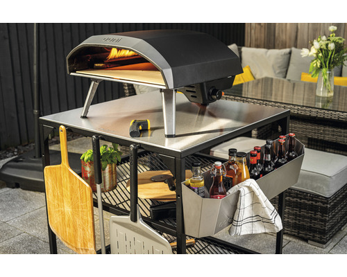 Ooni pizzaoven op een buitenkeukenwagen met accessoires in de tuin.