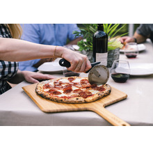 Scène van iemand die een pizza snijdt met een pizzasnijder op een houten plank, met een fles wijn en glazen op de achtergrond, op een tafel buitenshuis