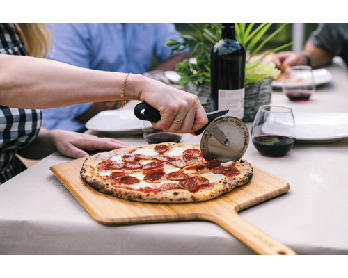 Scène van iemand die een pizza snijdt met een pizzasnijder op een houten plank, met een fles wijn en glazen op de achtergrond, op een tafel buitenshuis