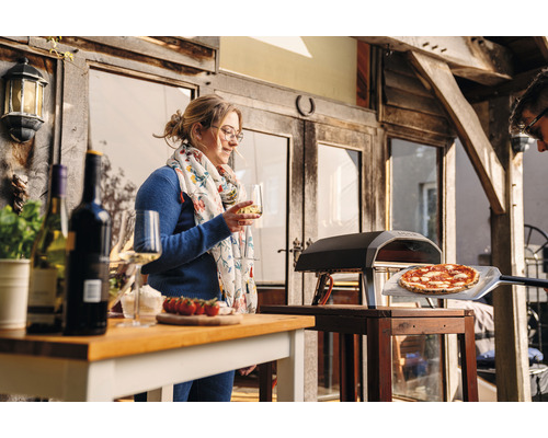 Scene met pizzaoven, pizza op een pizzaschep, tafel met flessen wijn, glazen en een vrouw met een glas wijn.