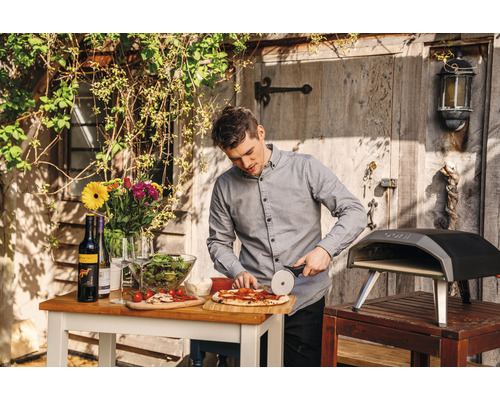 Man snijdt pizza in de tuin met pizzaoven, tafel, wijn, salade en bloemen.