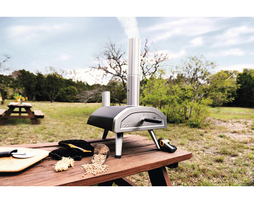 Ooni pizzaoven op een houten tafel in de buitenlucht met pizza-accessoires.