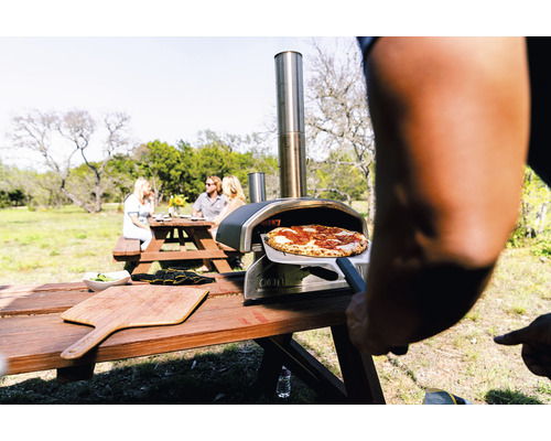 Buitenscène met een pizzaoven, een pizza op een pizzaschep en een groep mensen aan een picknicktafel.
