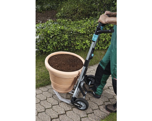 Een persoon vervoert een bloempot met een steekwagen in de tuin.