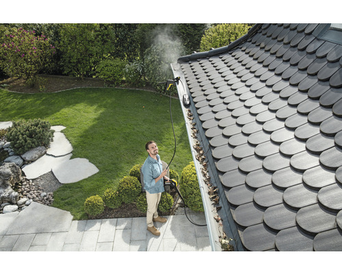 Een man maakt een dakgoot schoon met een hogedrukreiniger in een tuin.