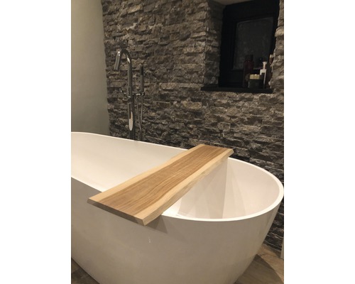 Vrijstaand bad met houten plank in een badkamer met een stenen muur.