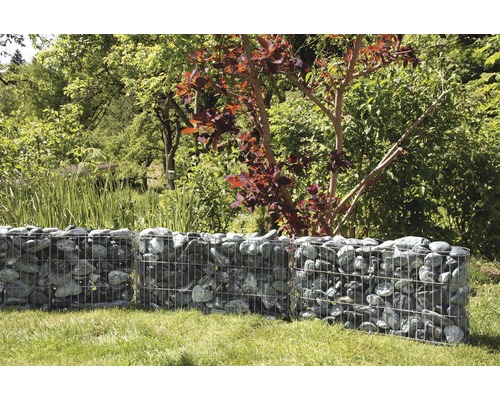 Schanskorfmuur in de tuin met bomen op de achtergrond.