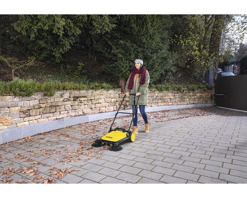 Vrouw gebruikt een gele veegmachine om bladeren van een oprit te verwijderen.