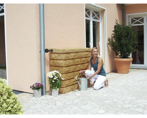 Scene met een vrouw die naast een regenwatertank knielt, met bloemen in potten en een gieter in een tuin.