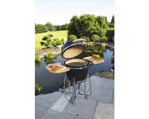 Keramische barbecue met open deksel, houten planken en roestvrijstalen frame in een tuinscène