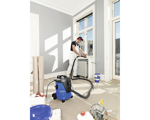 Ambachtsman schuurt met een schuurmachine een raam dat is aangesloten op een stofzuiger, in een lichte kamer.