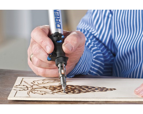 Een persoon gebruikt een Dremel brandgereedschap om een druivenmotief in een houten plank te branden.