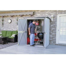 Een persoon opent een tuinhuis met een barbecue en tuingereedschap op een terras.