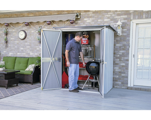 Een persoon opent een tuinhuis met een barbecue en tuingereedschap op een terras.