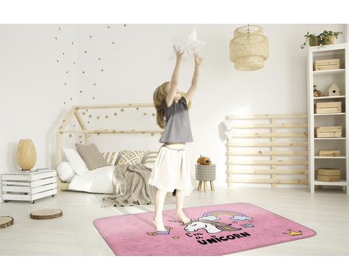 Een meisje springt op een rechthoekig, roze vloerkleed met eenhoornmotief in een kinderkamer.