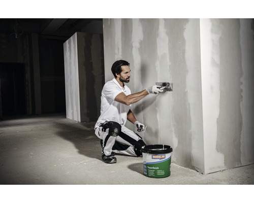 Een man egaliseert een muur met vulmiddel, terwijl een emmer vulmiddel op de grond staat.