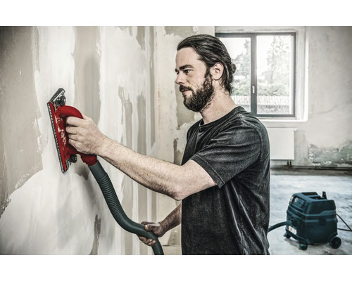 Ambachtsman schuurt een muur met een handschuurmachine aangesloten op een stofzuiger.