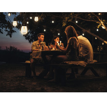 Groep mensen zit aan tafel onder een lichtslinger in de tuin.