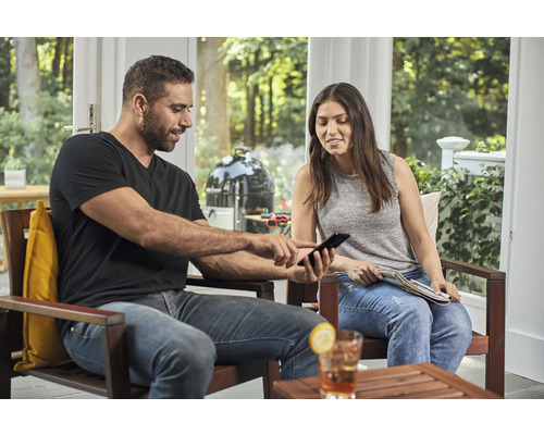 Man en vrouw zitten op het terras en kijken naar een mobiele telefoon, op de achtergrond een kogelbarbecue.