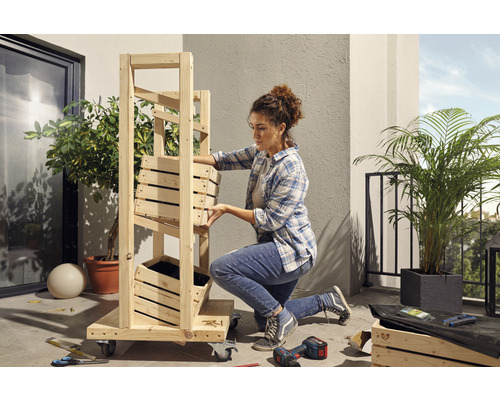 Een vrouw monteert een houten rek met plantenbakken op een balkon. Gereedschap en planten zijn ook zichtbaar.