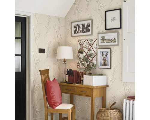 Hoekweergave van een kamer met bureau, stoel en decoratieve afbeeldingen aan de muur, behangen met een patroon van kleine bloemen en takken.