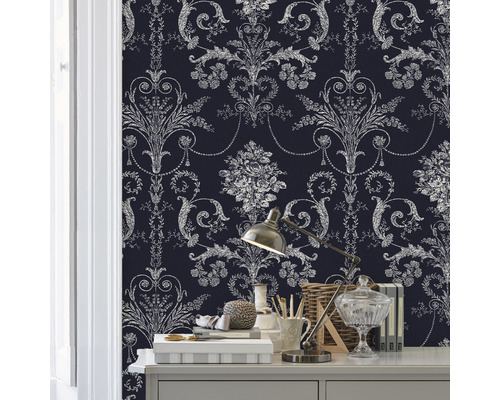 Close-up van een kamer met een muur bedekt met een behang met patroon. Op een tafel staan diverse decoratieve objecten en een lamp.