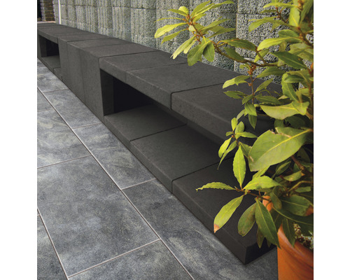 Lange, donkere tuinbank van betonstenen naast een groene struik en tegelvloer.