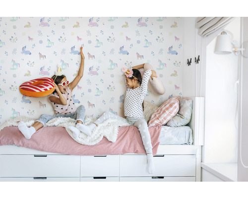 Twee meisjes spelen op een bed voor een behang met eenhoornmotief.