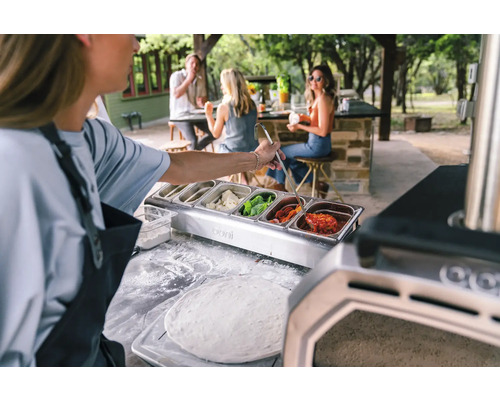 Persoon bereidt buiten pizza met ingrediënten uit roestvrijstalen containers.